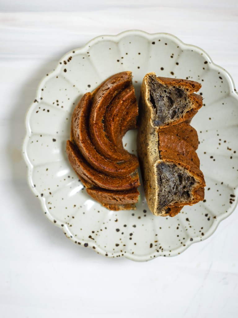 an image of mini black sesame mochi bundt cake, cut in half, on a off-white speckled scalloped shaped plate, with a marble-like surface